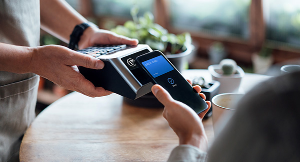 Close up of a male's hand paying bill with credit card contactless payment on smartphone in a cafe, scanning on a card machine.