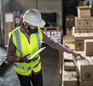 Warehouse worker wearing face mask and protective workwear checking products using digital tablet.