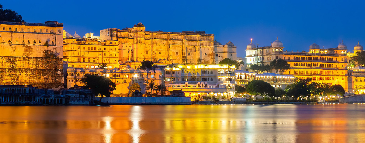 Evening view of illuminated Udaipur Palace in India with lights reflected in the water.