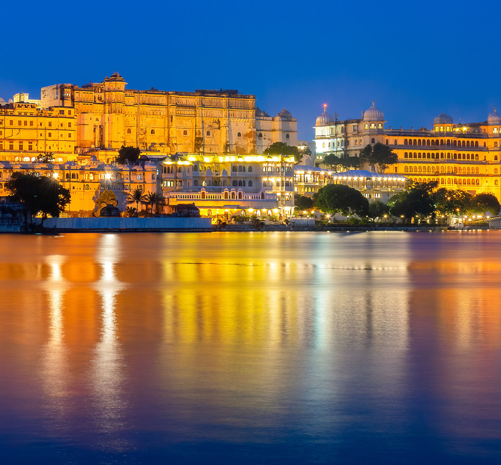 Evening view of illuminated Udaipur Palace in India with lights reflected in the water.