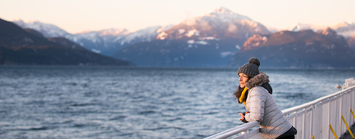 A woman looking out over the ocean during a sunset as she leans on a railing. She is wearing winter clothing and there are mountains off in the distance behind her.
