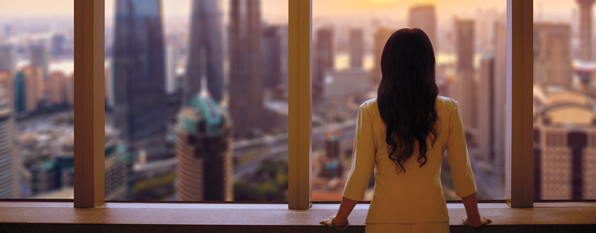 Woman in suit looks out at Shanghai skyline at sunset from window in building.