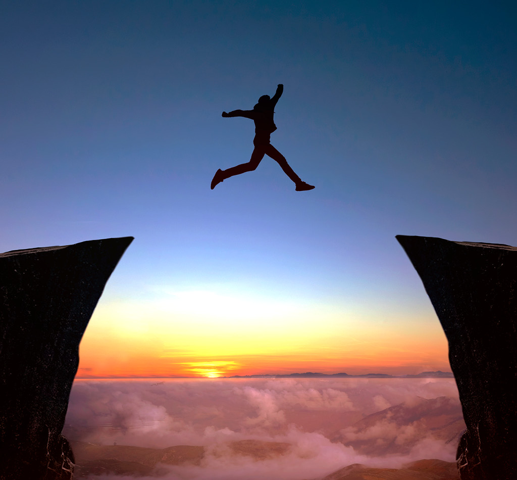 A person jumping from one steep cliff to another over a sunset sky.