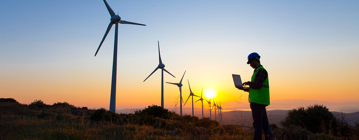 Image of wind turbines and a man holding an open laptop standing in front of a sunset/sunrise