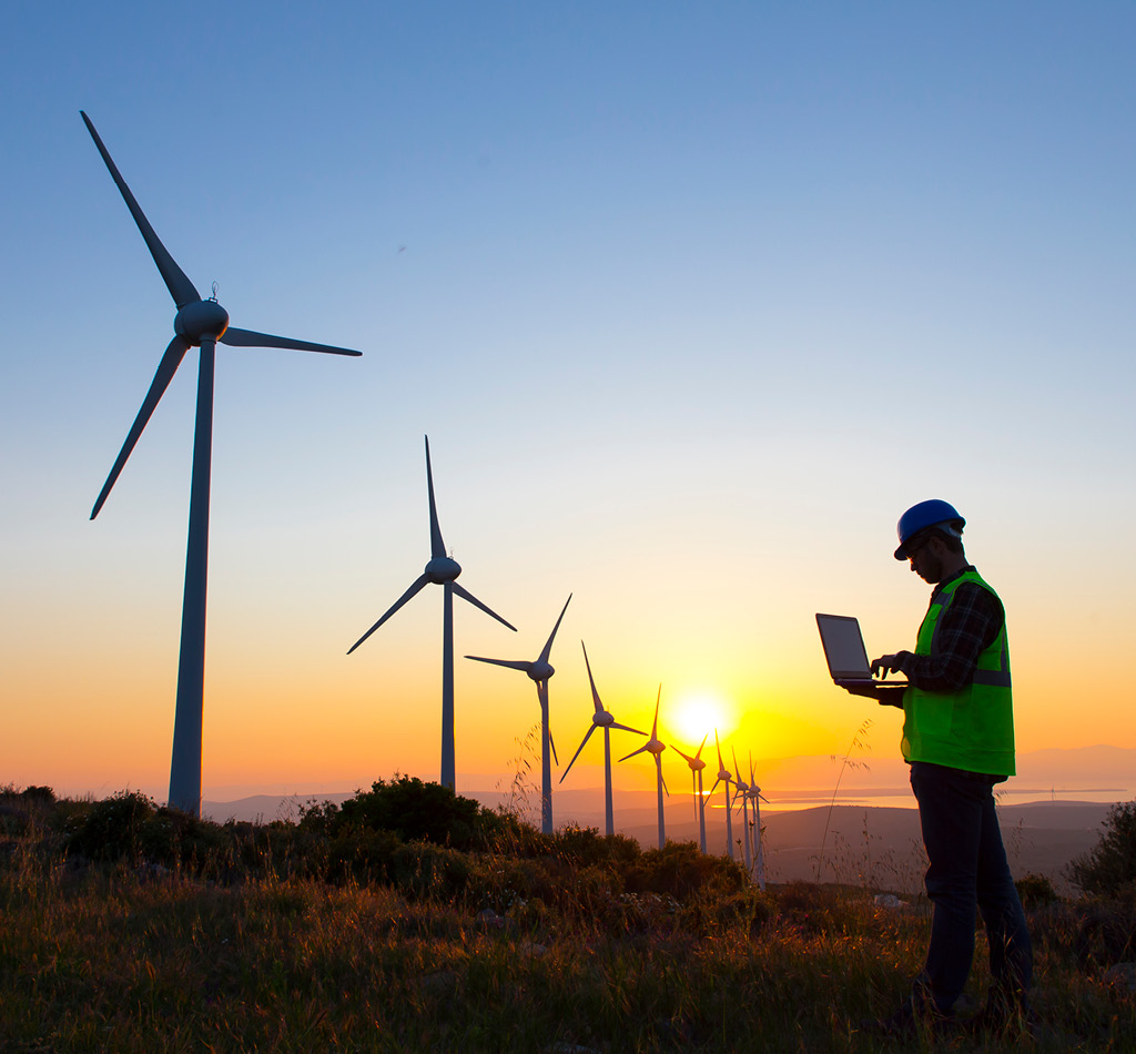 Image of wind turbines and a man holding an open laptop standing in front of a sunset/sunrise