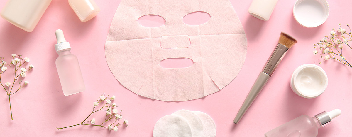 Facial sheet mask with different cosmetic products and flowers on pink background. 
