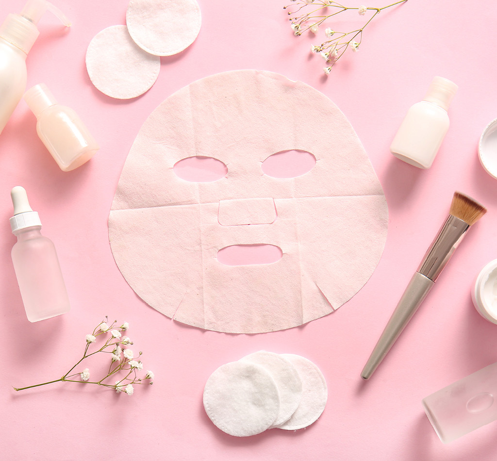 Facial sheet mask with different cosmetic products and flowers on pink background.