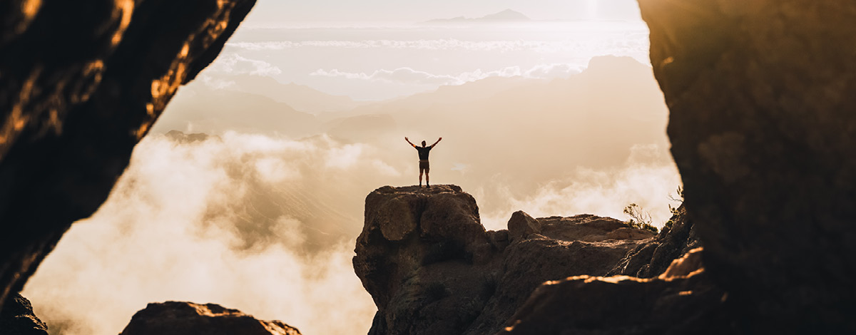 Man standing on the top of a high cliff during the sunset with raised hands.