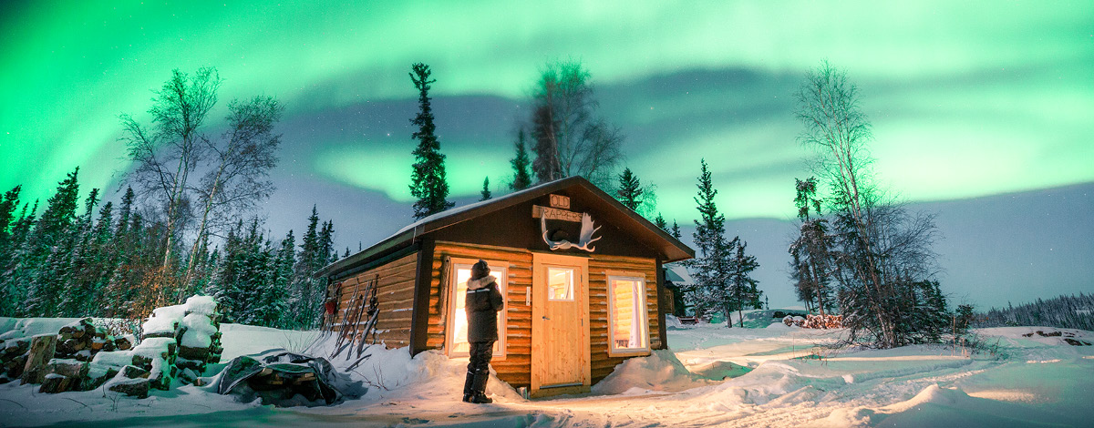 A traveller standing outside a cabin looking at the northern lights in Yellowknife, Northwest Territories, Canada.