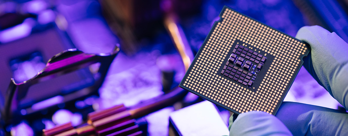 A hand holding a computer microchip with a motherboard in the background.