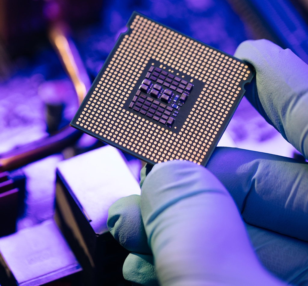 A hand holding a computer microchip with a motherboard in the background.