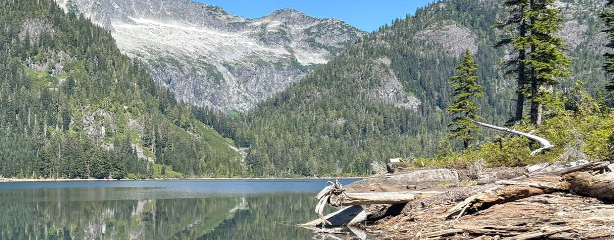 A clear lake reflecting surrounding trees and mountains under a bright blue sky.