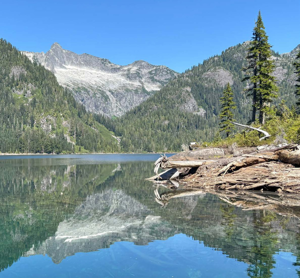 A clear lake reflecting surrounding trees and mountains under a bright blue sky.